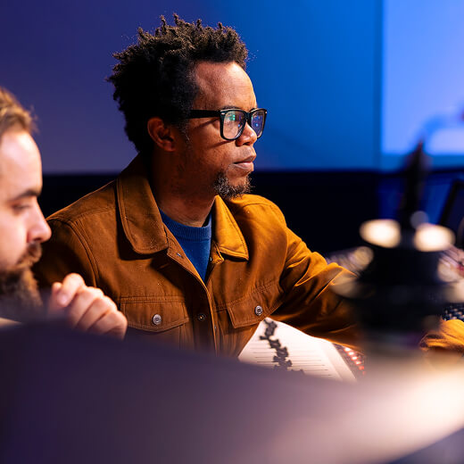 Two men sit at a mixing console in a dimly lit studio, focused on audio equipment, with one wearing glasses and a brown jacket.