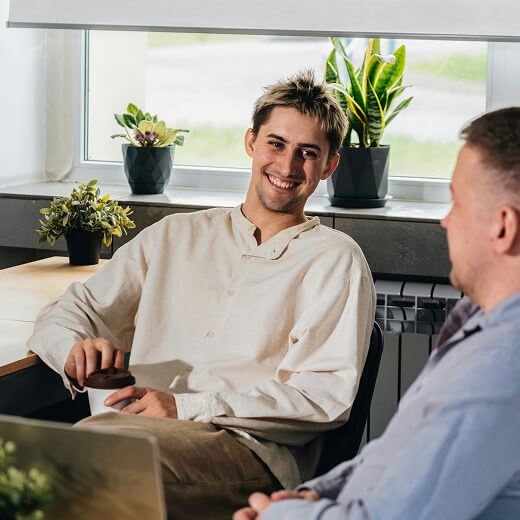 Two men sit and talk in a bright office space with potted plants on the windowsill; one man holds a coffee cup and smiles.