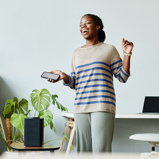 A woman smiling and holding a smartphone stands in a modern room with a speaker, a table, a laptop, and a potted plant in the background.