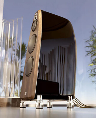A modern, high-end speaker with a glossy black finish and wooden sides sits on a clear platform indoors, with plants and sunlight visible in the background.