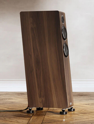 A tall, wooden floor-standing speaker with three visible drivers, placed on a parquet floor against a light-colored wall.
