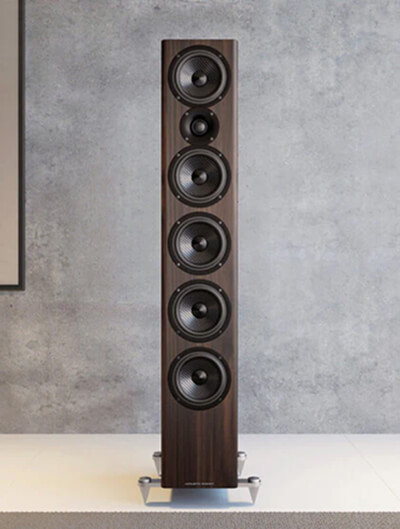 A tall, wooden floor-standing speaker with six circular drivers is positioned against a gray wall on a white base.